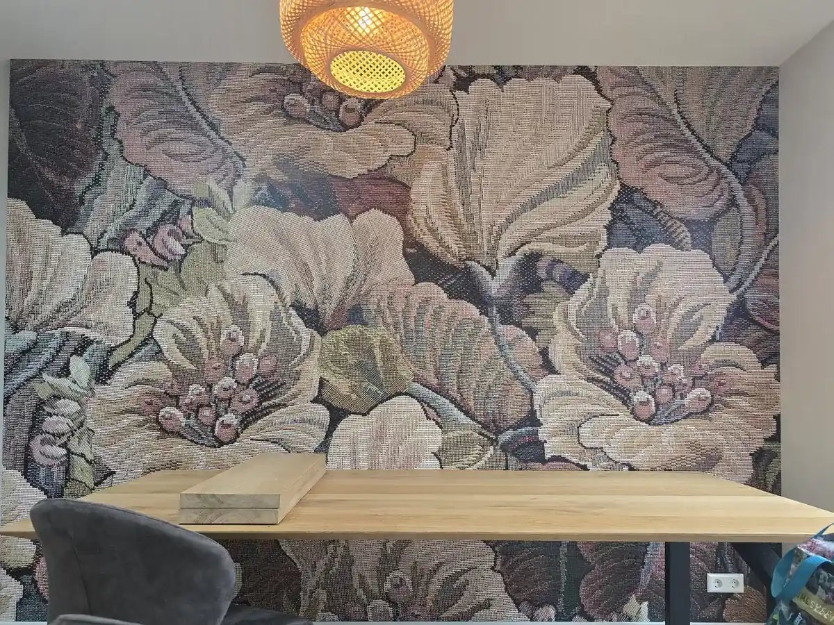 Eetkamer met een wand van vliesbehang met grote bloemen in oudroze, groen en crème, boven een houten tafel met een rieten hanglamp.