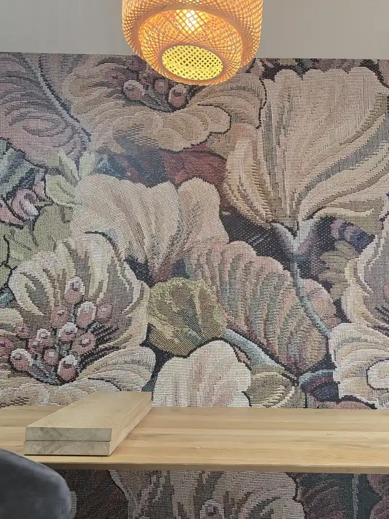Wand met vliesbehang met grote bloemen in oudroze, groen en crème, onder een rieten hanglamp en boven een houten tafel.