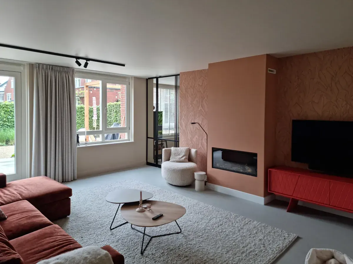 Lichte woonkamer met een terracotta schouw en wanden met vliesbehang in een bladmotief, een roestrode bank en een groot raam naar de tuin.