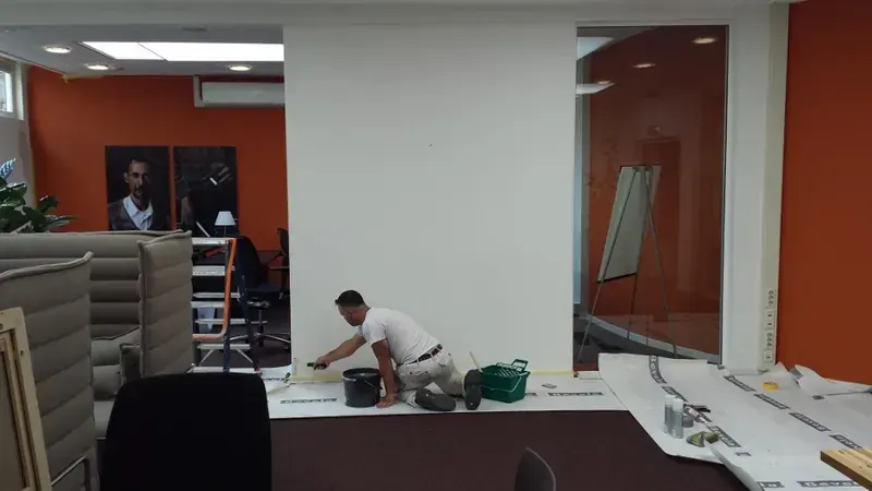 Schilder in een wit T-shirt latext een wand in een kantoor; de vloer is afgedekt en de oranje wand ernaast is al klaar.