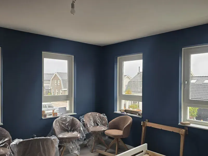Kamer in een nieuwbouwwoning met donkerblauw geschilderde wanden rond drie ramen; stoelen staan nog in plastic in het midden.