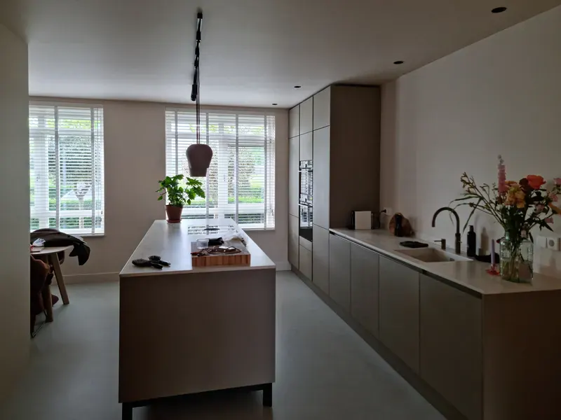 Lichte keuken met een lang kookeiland en strak afgewerkte witte wanden, met ramen naar de tuin.