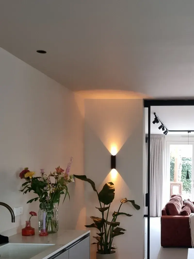 Strak afgewerkte witte wand met een zwarte wandlamp die het licht omhoog en omlaag werpt, naast een doorgang naar de woonkamer met een roestrode bank.